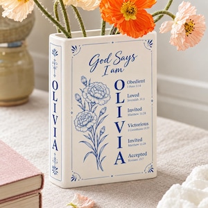 以下が含まれることがあります： 青い文字と花のイラストが描かれた、本型の白い陶器製花瓶。前面には「God Says I am OLIVIA」というテキストと、属性や聖書の節のリストが記されています。背表紙にも青い文字で「OLIVIA」と書かれています。