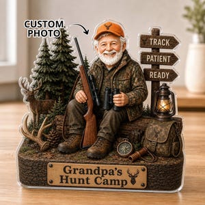 May include: A decorative figurine featuring a smiling older person in a camouflage jacket and orange cap, holding a rifle and binoculars. The base includes a sign reading 'Grandpa's Hunt Camp', a deer, pine trees, a lantern, and a signpost with 'TRACK', 'PATIENT', and 'READY'.