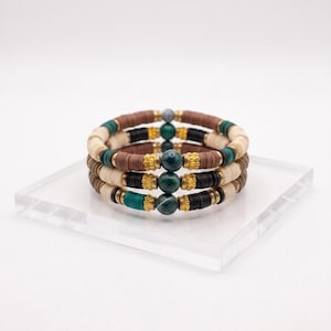 Op de afbeelding: Een stapel van drie armbanden met platte, ronde kleikralen in bruin, zwart, crème en blauwgroen. Elke armband bevat een centrale ronde groene steenkralen, geflankeerd door goudkleurige metalen tussenstukjes voor een gelaagd uiterlijk.