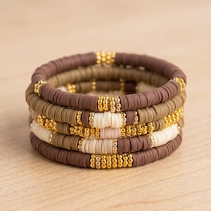 Op de afbeelding: Een stapel van vier armbanden gemaakt van kleine, platte, ronde kleikralen in tinten bruin, beige en crème. Elke armband is geaccentueerd met kleine, ronde, metallic gouden kralen die in groepjes over het ontwerp zijn verdeeld.