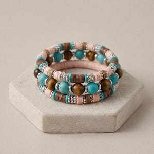 Op de afbeelding: Een stapel van drie armbanden met een combinatie van lichtroze kleischijfjes, donkerbruine houten kralen, turquoise ronde kralen, bruine tijgeroogkralen en kleine zilverkleurige metalen tussenstukjes in een herhalend patroon.