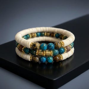 Op de afbeelding: Een stapel van drie armbanden met crèmekleurige platte schijfkralen, ronde blauwe en bruine gemarmerde stenen kralen en kleine goudkleurige metalen tussenstukjes. De middelste armband bevat drie cilindrische goudkleurige metalen kralen.