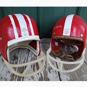 May include: Two vintage red football helmets with white centre stripes and cream-coloured plastic face masks. The left helmet features the Wilson brand name, while the right helmet displays a small graphic logo and interior leather padding.