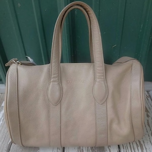 May include: A beige leather handbag with a textured surface, featuring two top handles and a zipper closure. The brand name Calvin Klein is embossed in the center of the front panel. The bag has a structured, rounded rectangular shape.