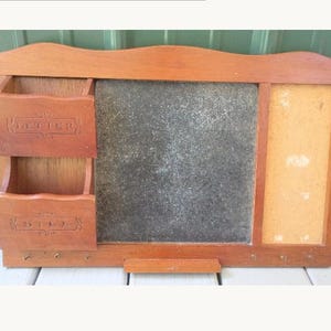 May include: A wooden wall organizer featuring two stacked storage compartments labeled "LETTER" and "BILL" on the left, a central rectangular chalkboard surface, and a vertical corkboard panel on the right, with small metal hooks along the bottom edge.