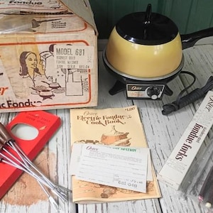 May include: An Oster Model 681 electric fondue pot in harvest gold with a black lid and handle, accompanied by its original cardboard box, a cookbook, a warranty card, and two sets of stainless steel fondue forks with brown handles in packaging.
