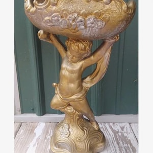 May include: A gold-colored decorative pedestal bowl featuring a sculpted figure of a cherub holding the basin aloft. The bowl has a textured, ornate rim and the base is decorated with floral motifs, all finished in a metallic gold paint.