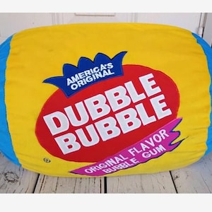 Puede incluir: Un cojín de peluche grande, suave y cilíndrico con forma de caramelo envuelto. El centro es amarillo con un óvalo rojo que presenta el texto blanco DUBBLE BUBBLE, AMERICA'S ORIGINAL y ORIGINAL FLAVOR BUBBLE GUM. Los extremos son azul brillante.