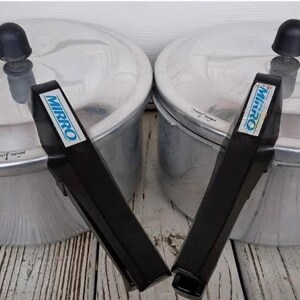May include: Two silver-colored aluminum pressure cookers with black plastic handles and locking lids. Each lid features a black knob and the brand name MIRRO printed in blue on the handle. The pots have a metallic finish and a circular design.
