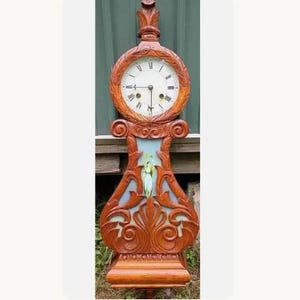 May include: A tall wooden wall clock featuring a circular face with black Roman numerals and two hands. The frame is intricately carved with scrollwork and leaf motifs. A central panel displays a painted figure of a person in a white dress.