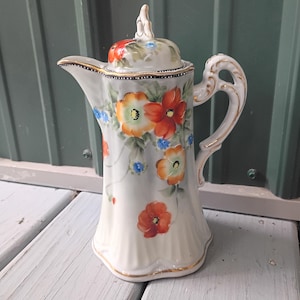 May include: A white porcelain coffee pot featuring a hand-painted floral pattern with orange, red, and blue flowers and green leaves. The pot has a scalloped base, a curved handle, a spout, and a matching lid with a decorative finial, all accented with gold trim.