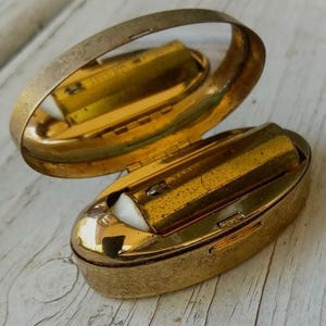 Puede incluir: Un estuche compacto ovalado de metal dorado con tapa abatible. El interior cuenta con un espejo en la tapa y un tubo de pintalabios dorado con base blanca encajado en la parte inferior. La superficie exterior tiene un acabado texturizado.