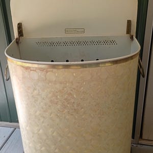 May include: A vintage Detecto New York metal hamper with a cream-colored finish and a textured diamond pattern on the exterior. The hamper features a hinged lid, a gold-toned metal rim, side handles, and a perforated ventilation strip near the top.