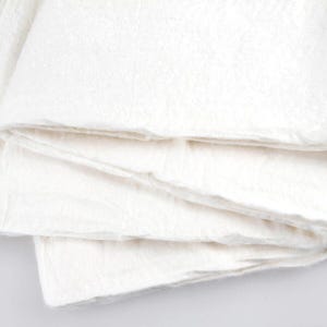 Op de afbeelding: Een stapel gevouwen, gebroken witte stof, mogelijk linnen of katoen, met een licht getextureerde uitstraling. De lagen zijn netjes gerangschikt, wat wijst op een strakke en eenvoudige esthetiek. De stof ziet er zacht uit.