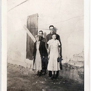 May include: A vintage black and white photograph of four people standing in front of a weathered building. Two women wear polka dot dresses, and the men are in dark shirts. The image has a grainy texture, typical of older photographs.