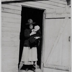 Old Photo Woman Holding Baby Wrapped in Blanket Woman wearing Dress Sweater and Hat standing in Doorway 1910s Photograph Snapshot Vintage