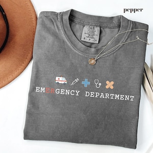 May include: A charcoal gray t-shirt featuring the text EMERGENCY DEPARTMENT in white letters with a red letter E. Above the text are small icons of an ambulance, a syringe, a blue cross, a stethoscope, and a bandage. The shirt has a crew neck.