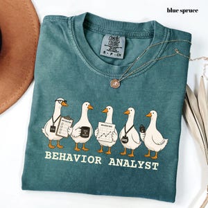 Op de afbeelding: Een blauw-sparrenkleurig t-shirt met een afbeelding van vijf witte ganzen. De ganzen zijn afgebeeld als gedragsanalisten die voorwerpen vasthouden zoals een klembord, een mok, een grafiek, een stopwatch en een clicker. Tekst BEHAVIOR ANALYST.