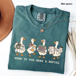 May include: A blue spruce coloured t-shirt featuring a graphic of four white geese dressed as pharmacists. The text reads "HONK IF YOU NEED A REFILL". The geese are holding a mortar and pestle, a pill bottle, a stack of books, and a clipboard.