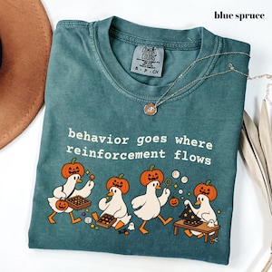 May include: A blue spruce short-sleeve t-shirt with a graphic print of four white ducks wearing pumpkin heads. The text reads, behaviour goes where reinforcement flows. The shirt features a Comfort Colors brand label at the inner neckline.