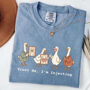 May include: A blue crew neck t-shirt featuring a graphic of four white geese. One goose wears a green jacket, one holds a framed picture, one has large pink lips and gives a thumbs up, and one wears a brown jacket. The text reads, Trust Me, I'm Injecting.