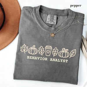 Könnte beinhalten: Ein pfefferfarbenes Kurzarm-T-Shirt mit einer hellen Grafik aus Blättern, Kürbissen und einem Kaffeebecher mit Herz sowie dem Text "BEHAVIOR ANALYST", der in einer fetten, serifenlosen Schrift auf der Brust aufgedruckt ist.