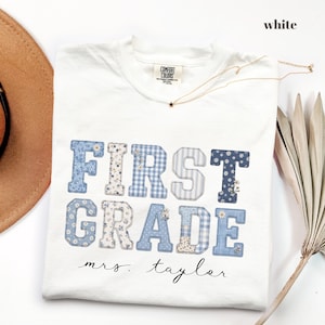 Może przedstawiać: Biały t-shirt z krótkim rękawem z napisem FIRST GRADE wykonanym niebiesko-białymi wzorzystymi literami blokowymi w kwiaty i kratkę. Poniżej tekstu nadrukowano imię mrs. taylor czarną czcionką imitującą pismo odręczne.