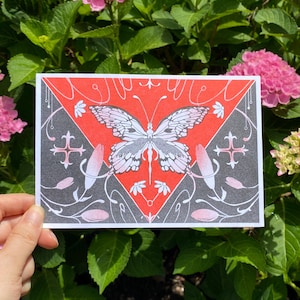 May include: A rectangular art print featuring a central white butterfly with detailed wings, set against a vibrant red triangular background. The surrounding area is dark gray with white floral motifs, stylized vines, and cross-like symbols.