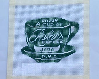 Op de afbeelding: Een vierkant naaldpunt canvas met een donkergroen kruissteekontwerp van een koffiekopje op een witte rasterachtergrond. De kop toont de tekst Enjoy A Cup Of, een cursief logo, Coffee, Java en N.Y.C. Het canvas heeft een crèmekleurige stoffen rand.