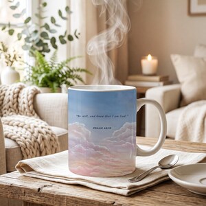 May include: A white ceramic mug featuring a printed design of a soft blue sky with pink and white clouds. The text "Be still, and know that I am God." and "PSALM 46:10" is printed in dark blue on the front of the mug, which has a curved handle.