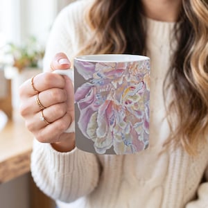May include: A white ceramic mug featuring a detailed, painterly illustration of a large, blooming flower with soft, layered petals in shades of pale pink, cream, and light blue, set against a solid, muted taupe-colored background.