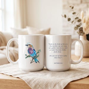 May include: Two white ceramic mugs. One mug features a colorful illustration of a lilac-breasted roller bird perched on a branch. The other mug displays the text: "Look at the birds of the air: they neither sow nor reap nor gather into barns, and yet your heavenly Father feeds them. Are you not of more value than they?" followed by "MATTHEW 6:26".