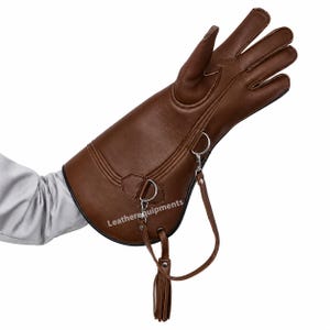 Könnte beinhalten: Ein brauner Lederhandschuh mit langer Stulpe, silberfarbenen Metallringen und einem abnehmbaren Lederriemen mit Quaste. Der Text "Leatherequipments" ist in Weiß auf der Oberfläche der braunen Lederstulpe aufgedruckt.