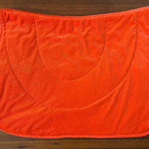 May include: A bright orange horse saddle pad featuring a quilted texture with a subtle, repeating dot pattern across the surface. The pad has a curved, contoured shape designed for a saddle, with matching orange piping along the outer edges.