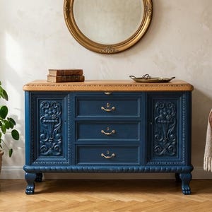 Könnte beinhalten: Ein marineblaues Sideboard aus Holz mit drei mittigen Schubladen mit messingfarbenen Griffen, flankiert von zwei Schranktüren mit geschnitzten floralen Reliefpaneelen. Das Möbelstück hat eine naturfarbene Holzplatte und steht auf vier geschnitzten Füßen.