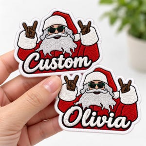 May include: Two embroidered patches featuring a cartoon Santa Claus wearing green sunglasses and making rock-and-roll hand gestures. One patch displays the word Custom in white script, while the other displays the name Olivia in white script.