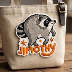 May include: A beige canvas tote bag featuring an embroidered patch of a gray and black raccoon. The patch includes orange star accents and the text JIMOTHY in orange lettering. A dark brown leather strap with a gold buckle is attached to the bag.