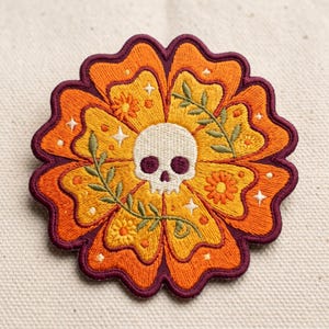May include: A circular embroidered patch featuring a central white skull with dark eye sockets, surrounded by stylized orange and yellow flower petals. The design includes small green leafy sprigs, tiny orange blossoms, and white four-pointed stars, all outlined with a thick dark purple border.