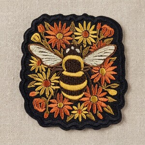 May include: A square embroidered patch with a scalloped black border featuring a central bumblebee with yellow and black stripes and white wings, surrounded by a dense arrangement of orange, yellow, and gold flowers on a black background.