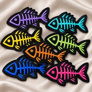 May include: A collection of seven embroidered fish skeleton patches in vibrant colors including purple, turquoise, yellow, lime green, orange, blue, and pink. Each patch features a black outline and a detailed skeletal structure design.