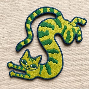 May include: An embroidered patch featuring a stylized cat with a long, curved body. The cat is lime green with dark green stripes, large almond-shaped eyes, and a dark blue border. The cat is depicted in a flexible, stretching pose.