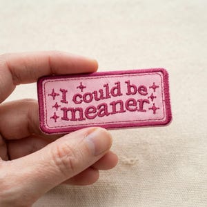 May include: A rectangular embroidered patch with a light pink background and a dark pink border. The text "I could be meaner" is stitched in dark pink lettering, accented by small four-pointed star symbols surrounding the words.