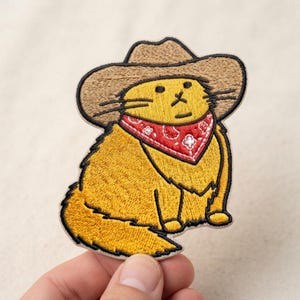 May include: An embroidered patch featuring a yellow cat wearing a tan cowboy hat and a red bandana with a white paisley pattern. The cat has a simple facial expression with black stitched eyes, nose, and mouth, and a long, fluffy tail.