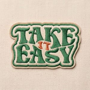 May include: An embroidered patch featuring the text TAKE IT EASY in dark green thread with the word IT in orange thread. The lettering is set against a cream-colored background with a matching cream-colored border that follows the shape.