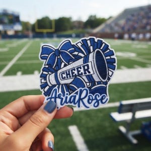 May include: An embroidered patch featuring a white megaphone with the word CHEER in dark blue letters, surrounded by dark blue pom-poms and a bow. The name Aria Rose is embroidered in dark blue script at the bottom of the patch.