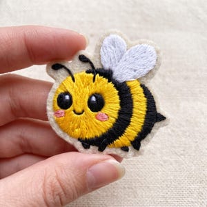 May include: A small, embroidered bee patch featuring a bright yellow and black striped body, large black eyes, pink cheeks, and white wings. The design is stitched onto a light cream-colored felt backing with a scalloped edge finish.