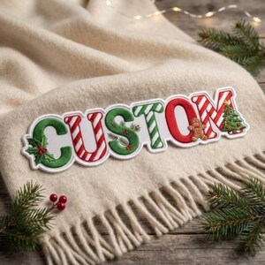 May include: A white fabric patch featuring the word CUSTOM in embroidered letters. The letters are decorated with green holly leaves, red berries, red and white candy cane stripes, pine branches, a gingerbread man, and a Christmas tree.