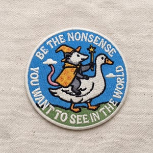 May include: A circular embroidered patch featuring a white opossum wearing a yellow wizard hat and cape, holding a star-tipped wand while riding a white goose. The blue background includes the text BE THE NONSENSE YOU WANT TO SEE IN THE WORLD.