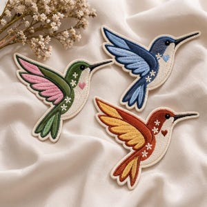 May include: Three embroidered hummingbird patches in green and pink, blue and white, and orange and yellow colorways. Each bird features a cream-colored chest, small white stitched flower accents, and a tiny heart detail on the breast area.