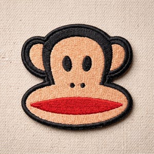 May include: An embroidered patch featuring a stylized monkey face with a tan complexion, black outlines, two black oval eyes, two small black nostrils, and a prominent wide red mouth. The design is symmetrical with rounded ears on both sides.